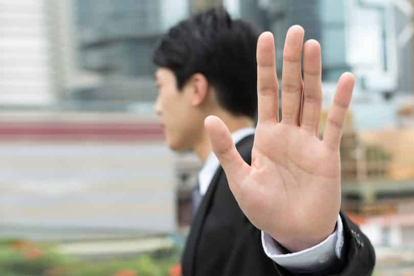 Homem na rua de roupas sociais estendendo a mão aberta como sinal de recusa.