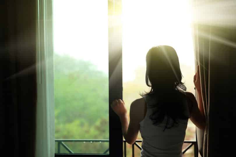 Mulher de costas abrindo a janela de sua sacada pela manhã.