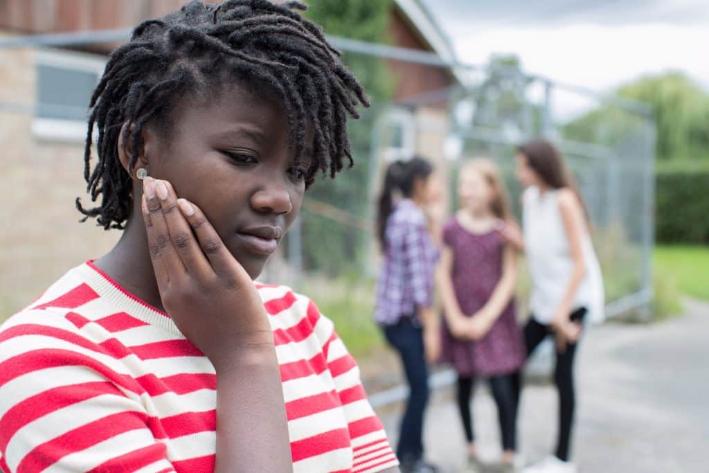 Menina negra vista de perfil, triste, enquanto outras meninas brancas estão reunidas atrás, rindo.