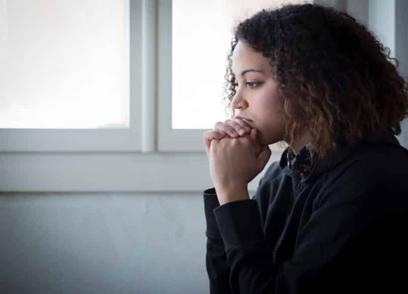 Mulher negra de cabelos cacheados vista de perfil, com semblante triste e as mãos sob o queixo.