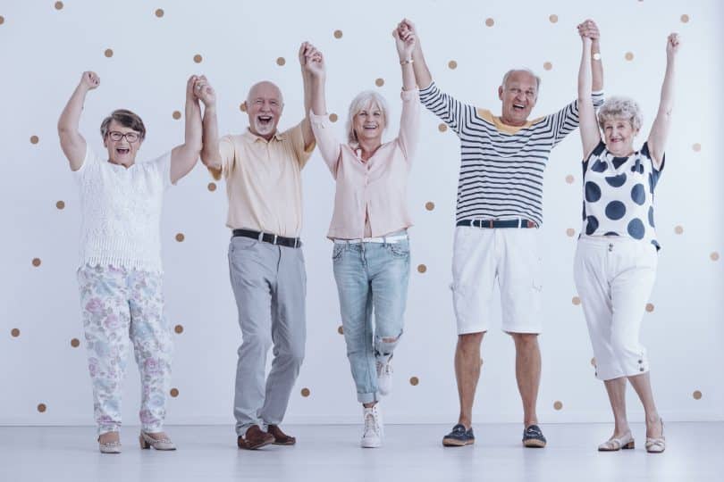 Grupo de cinco idosos sorrindo com as mãos para cima.