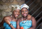 Imagem de duas amigas afrodescendentes. Elas estão alegres. Usam um corpete nas cores azul e branco e um turbante em suas cabeças, além de colares coloridos.