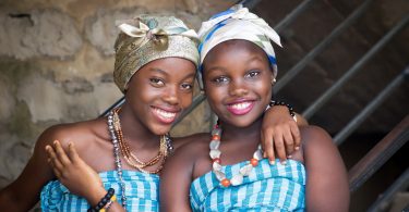 Imagem de duas amigas afrodescendentes. Elas estão alegres. Usam um corpete nas cores azul e branco e um turbante em suas cabeças, além de colares coloridos.