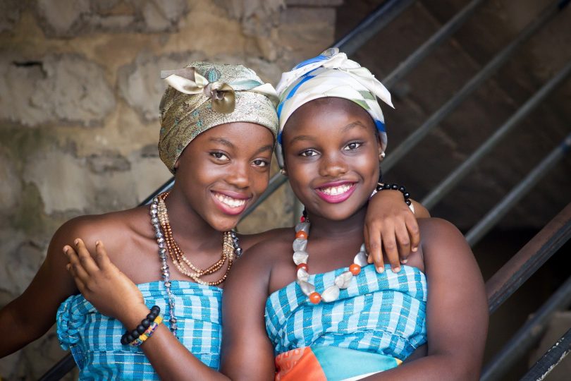 Imagem de duas amigas afrodescendentes. Elas estão alegres. Usam um corpete nas cores azul e branco e um turbante em suas cabeças, além de colares coloridos.