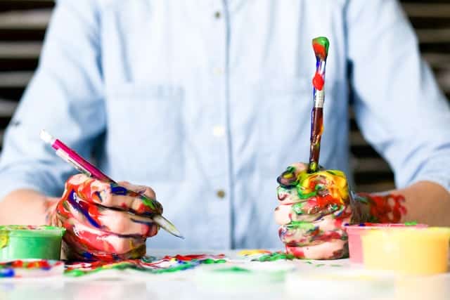 Homem com mãos sujas de tintas coloridas segurando caneta e pincel