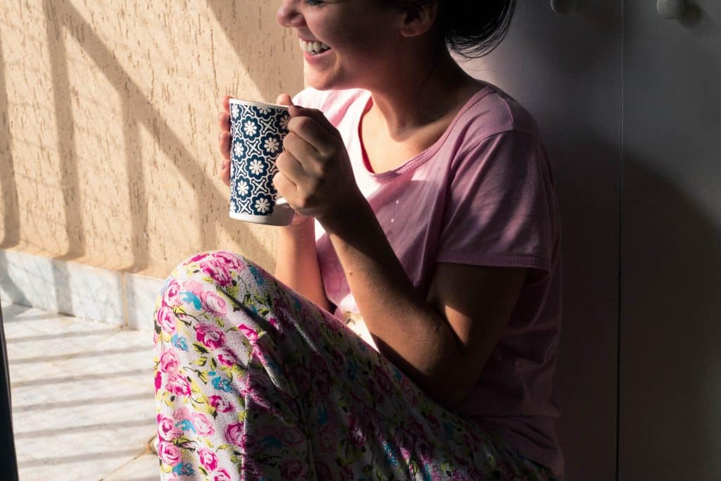 Mulher sentada na cama olhando para o lado sorrindo e segurando sua xícara