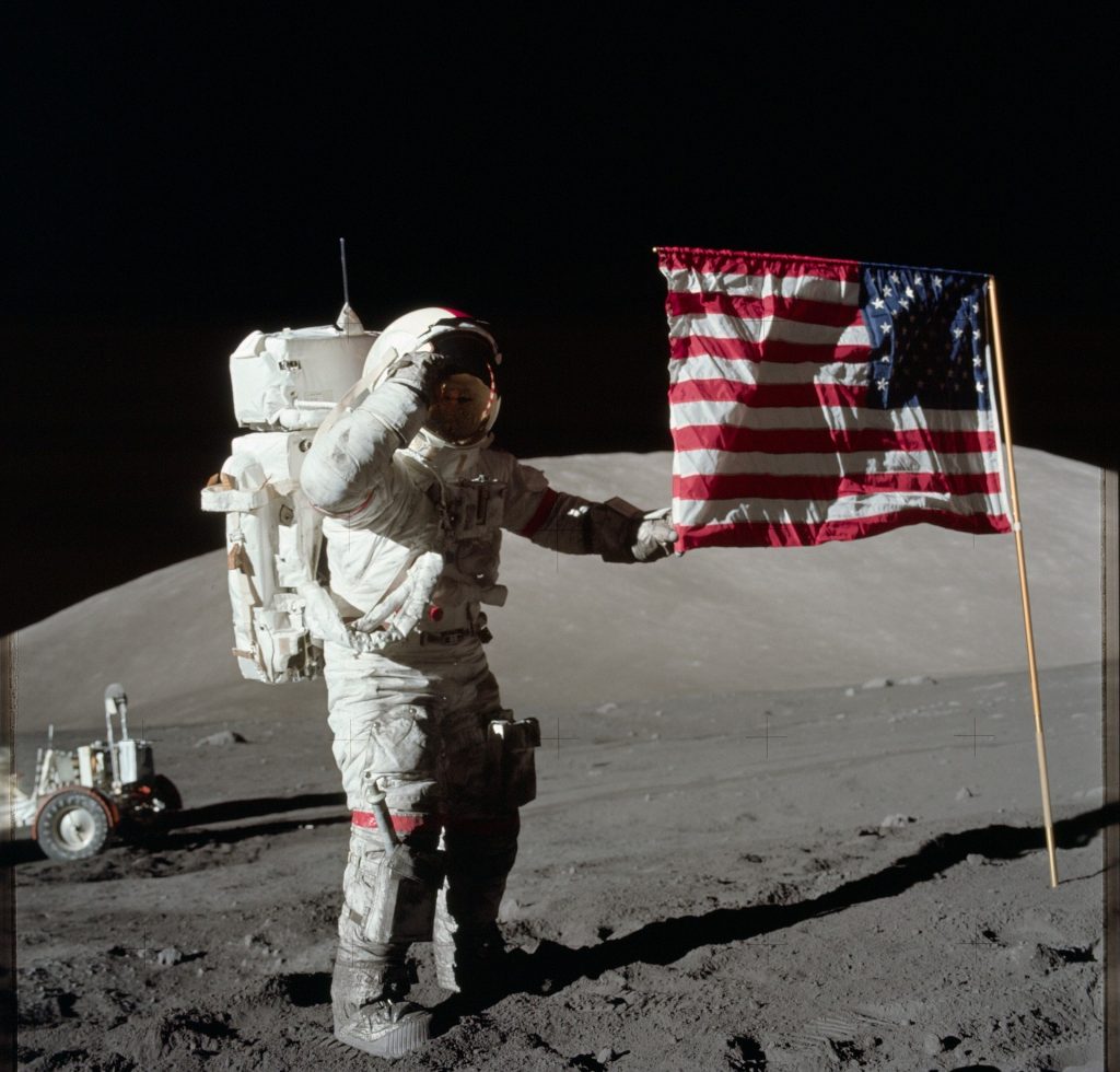 Imagem do primeiro homem astronauta pisando sobre a lua. Ao lado dela a bandeira dos Estados Unidos.
