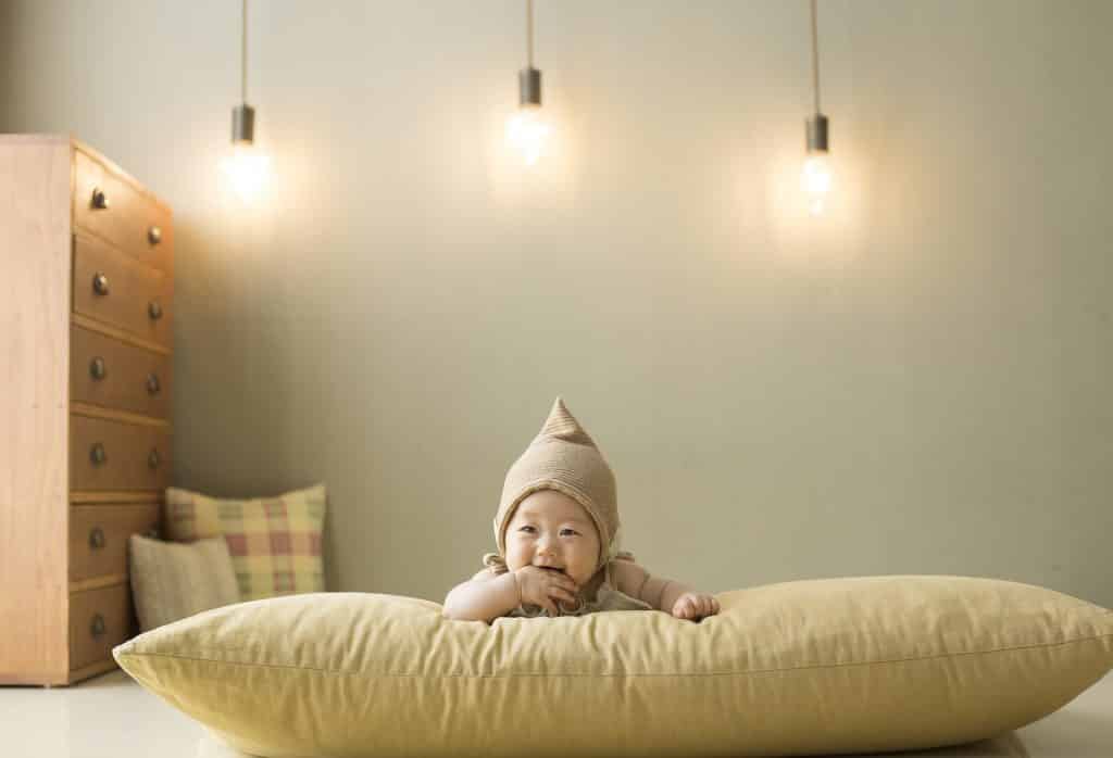 Imagem de um lindo bebê masculino oriental deitado de bruço sobre um grande almofadão disposto no chão do seu quarto.
