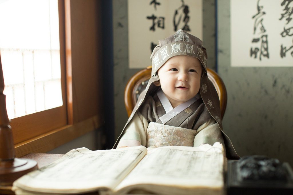 Imagem de um bebê coreado, usando uma roupa da cultura sentado em uma cadeirinha.