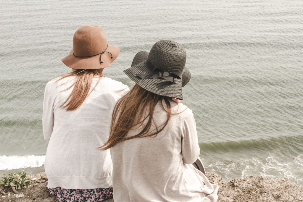 Duas mulheres sentadas à beira do mar. Elas estão de costas olhando para ele. Ambas usam um chapéu de sol e usam uma blusa de linha leve.