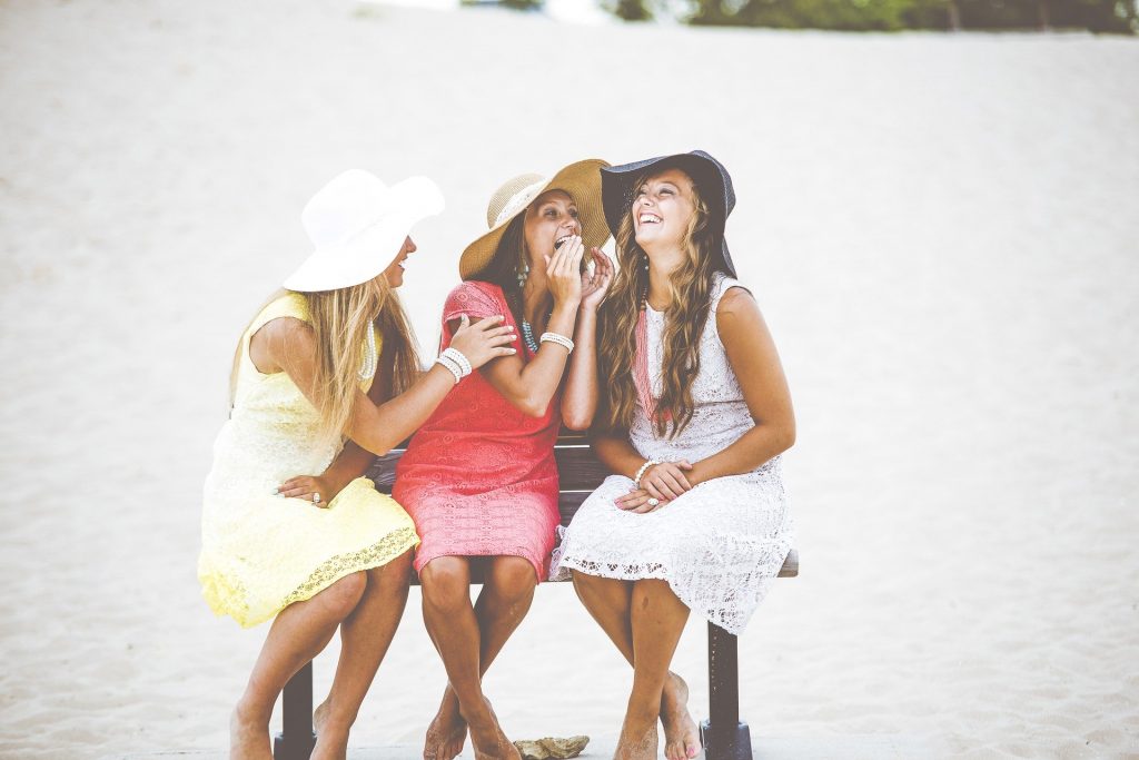 Três lindas e felizes amigas sentadas em um banco a conversar. Elas usam chapéus e vestidos de renda. Ela com a autoestima elevada e de bem com a vida.