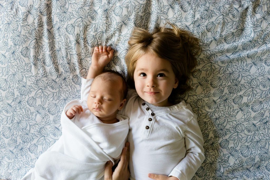 Imagem de dois irmãos, sendoo um recém nascido e uma maior, deitados sobre a cama.
