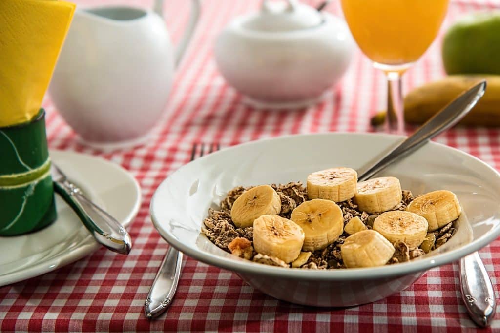 Imagem de um prato de cereais com bananas picadas. Ele está servido sobre uma mesa com toalha quadriculada nas cores vermelho e branco. Ao lado do prato um copo de suco de laranja.
