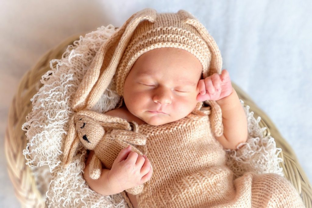 Imagem de um bebê vestido com uma roupa de coelho de lã, deitado em um cesto de palha. Ele está dormindo e segura em sua mão um coelho de lã.