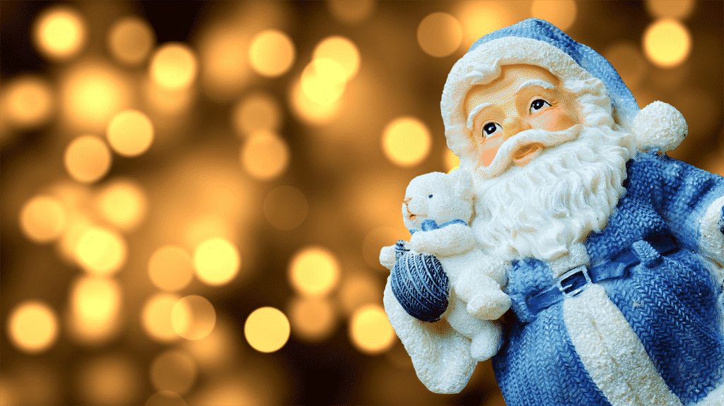 Imagem de um lindo boneco de papai noel vestindo uma roupa azul e segurando em uma de suas mãos um ursinho de pelúcia branco.