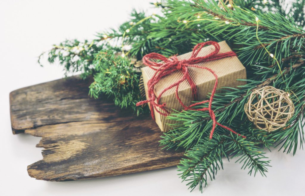 Imagem de um presente de Natal em uma caixa de papelão pequena embrulhada com um laço de linha na cor vermelho. A caixa está sobre uma madeira decorada com folhas da árvore de Natal.