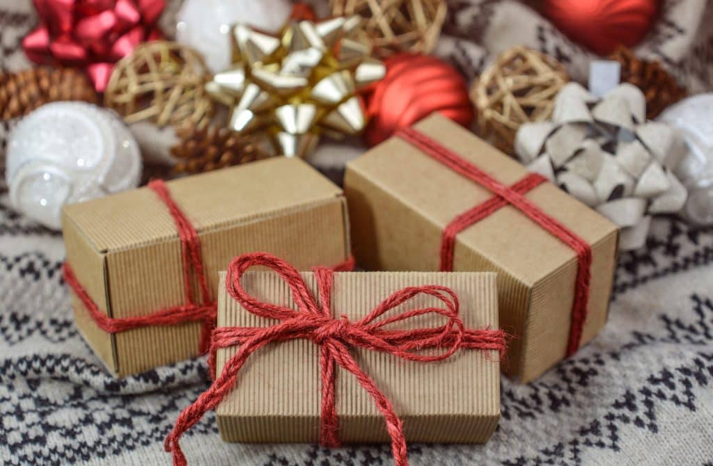 Imagem de três caixas pequenas de papelão de presente de Natal embrulhadas com uma linha vermelha e um laço, sobre uma mesa com toalha de renda branca e enfeites natalinos.