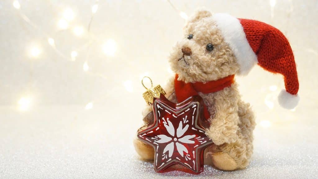 Imagem de  um lindo e pequeno ursinho de pelúcia vestindo uma touca de papai noel e segurando em suas mãos um pingente em formato de estrela vermelha para colocar na árvore.
