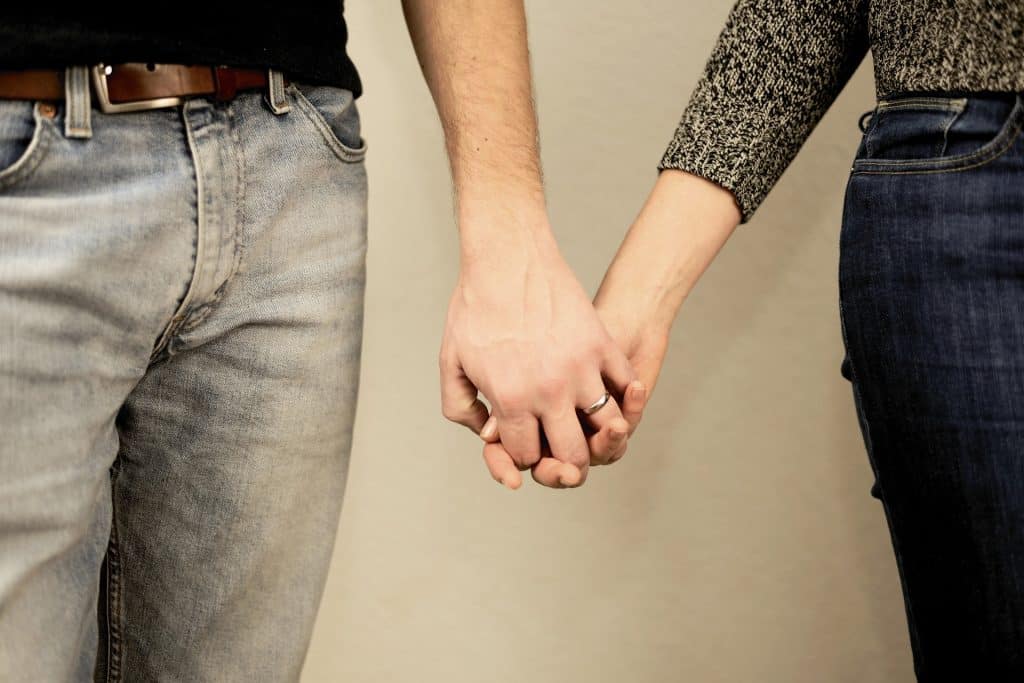 Imagem de um casal de homem e mulher - mostrando do pescoço para baixo. Eles estão lado a lado e de mãos dadas.