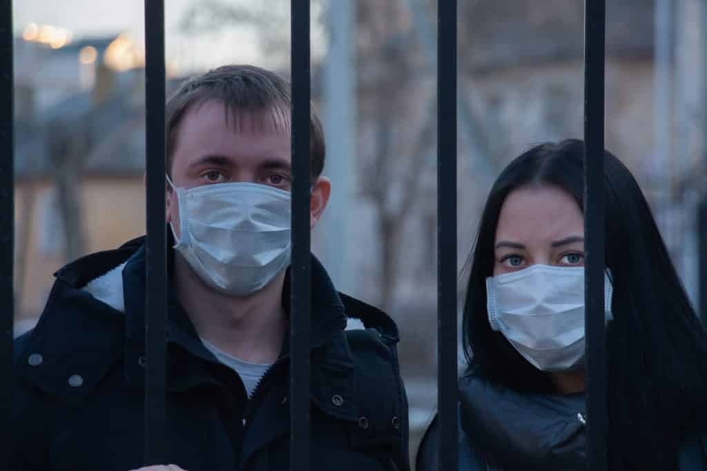 Imagem dos rostos de um casal de homem e mulher atrás de um portão proibidos de sair devido ao novo Coronavírus. Ambos usam máscara.