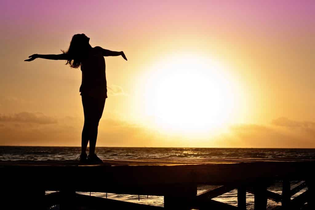 Silhueta de uma mulher de braços abertos ao pôr do sol.