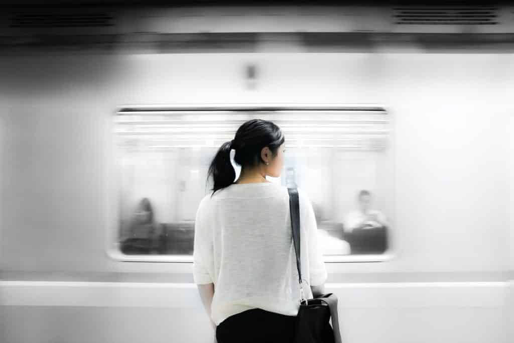 Mulher em pé, vista de costas, olhando para o lado enquanto um metrô passa a sua frente.