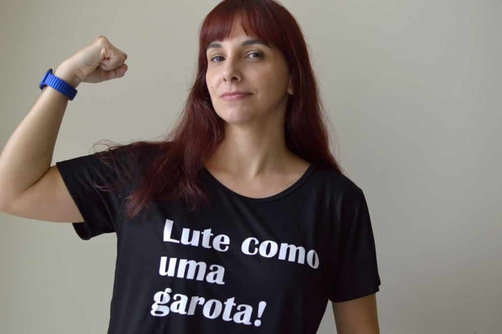 Imagem de uma mulher de cabelos longos avermelhados. Ela usa uma camise preta com os dizeres: Lute como uma garota!
