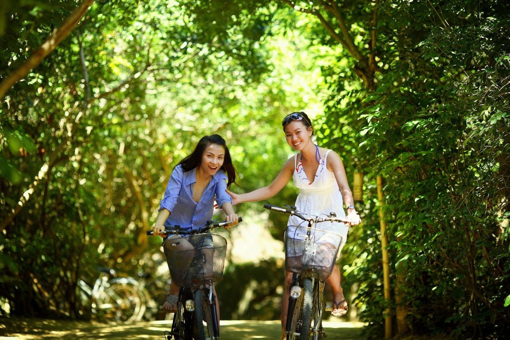 Imagem de duas amigas orientais andando de bicicleta no meio de uma floresta. Elas estão alegres e conseguiram um tempo para estarem juntas e fazer o que mais gostam.
