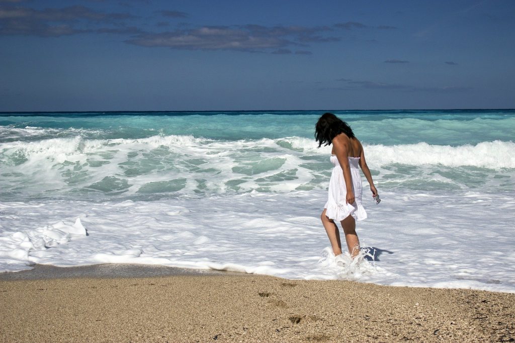 Imagem de um garota usado um vestido branco. Ela está caminhando na praia próximo ao mar. Ela aparenta está feliz, sozinha e com a autoestima elevada.
