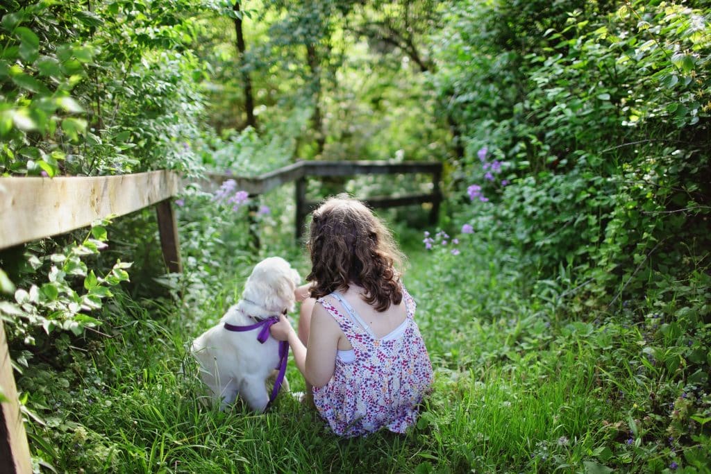 Menina branca de costas para a câmera em meio à plantas com um cachorro.