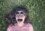 Imagem divertida de uma mulher deitada sobre um gramado. Ela usa um óculos gigante, um bigodão e uma sobrancelha enorme. Ela está muito alegre e com a autoestima elevada.