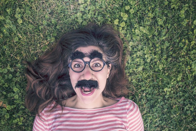 Imagem divertida de uma mulher deitada sobre um gramado. Ela usa um óculos gigante, um bigodão e uma sobrancelha enorme. Ela está muito alegre e com a autoestima elevada.