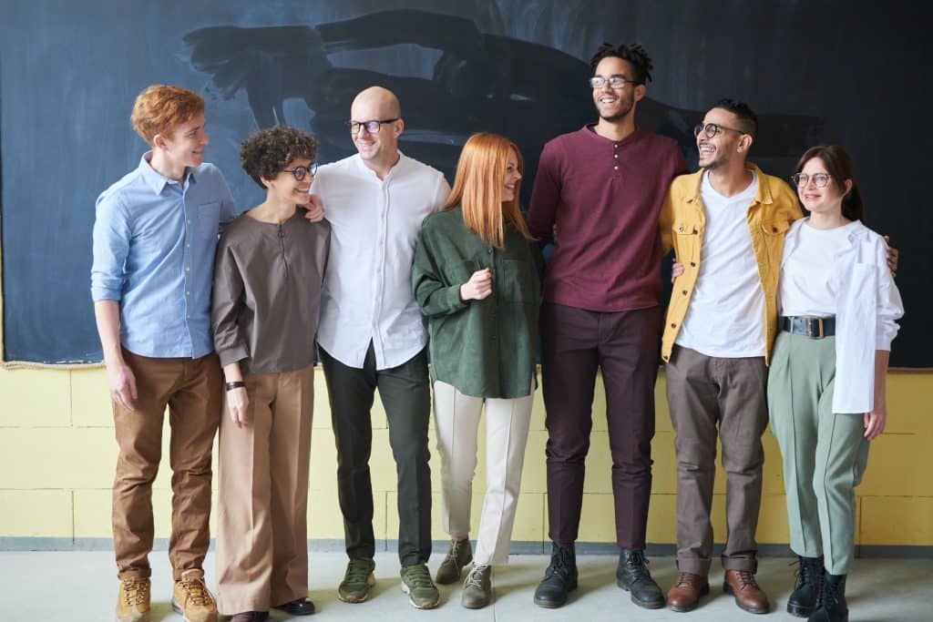 Grupo de pessoas de diferentes idades, cores e etnias em pé, em frente a um quadro negro.