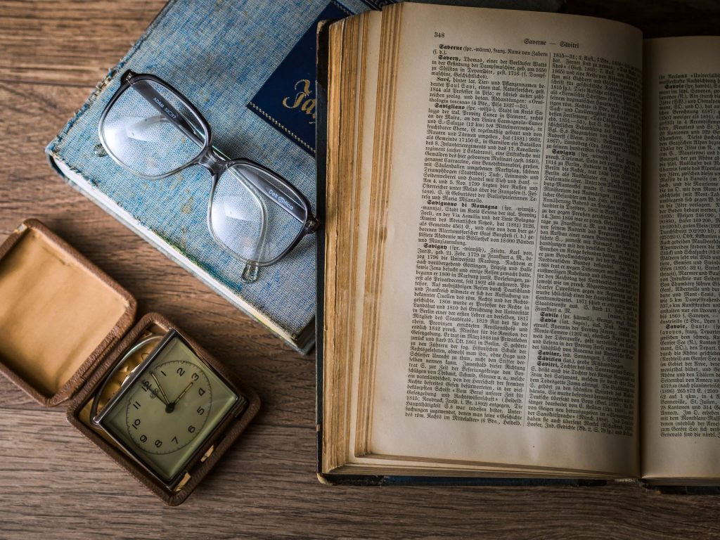 Imagem do livro de Kepler aberto sobre uma mesa. Ao lado dele um óculo de grau, uma agenda de capa azul e um relógio antigo.