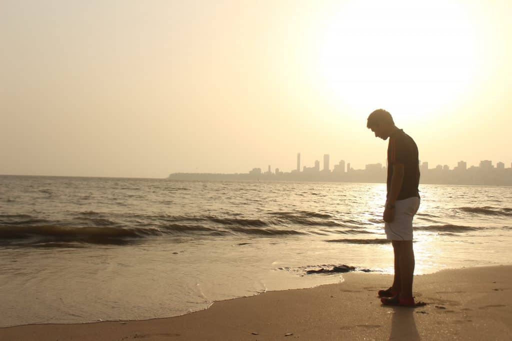 Homem em pé na praia, em frente ao mar, com a cabeça baixa.