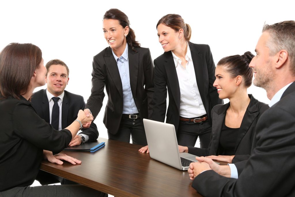 Imagem de mulheres e homens e uma mesa de negociação em um ambiente corporativos. Ambos, mulheres e homens vestem ternos e camisas, representando a igualdade de gênero.