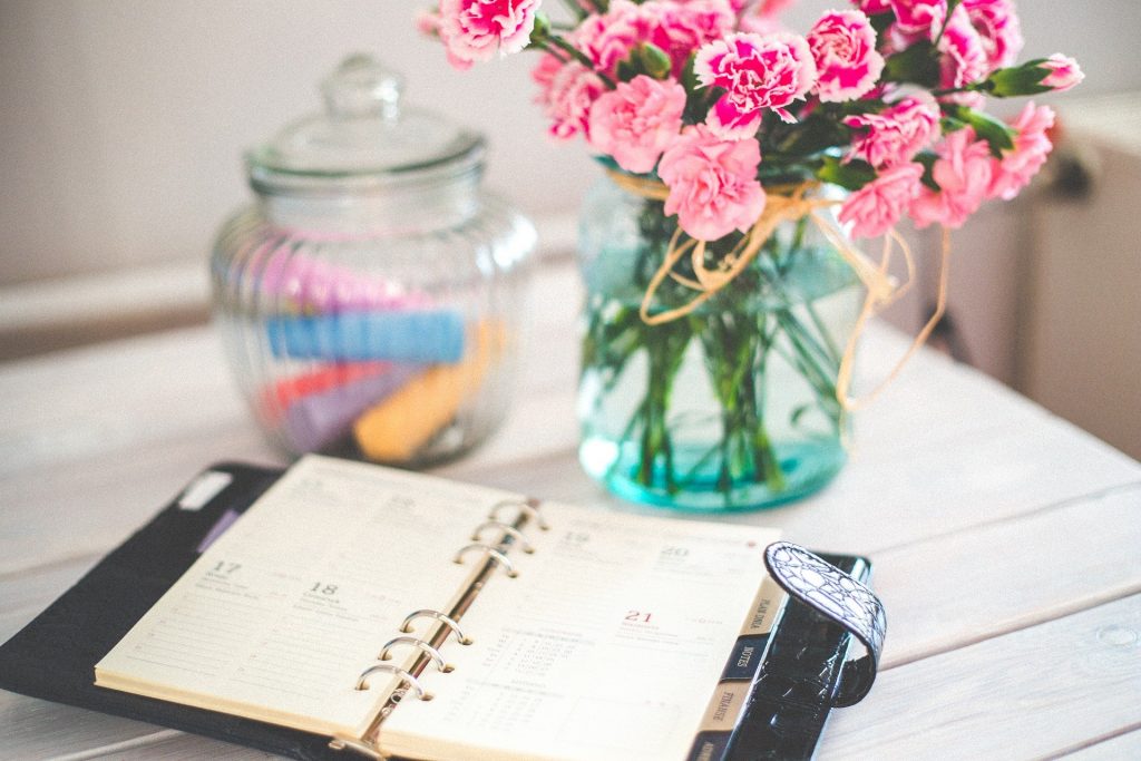 Imagem de uma agenda sobre uma mesa para preparar o planejamento para o ano novo. Ao lado um vaso de flores rosas e um pote com giz colorido.