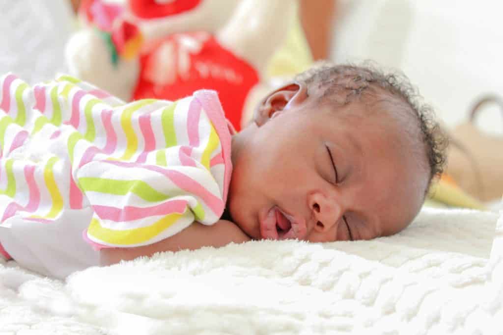 Imagem de um lindo bebê masculino da cor negra deitado de bruços sobre a sua cama.
