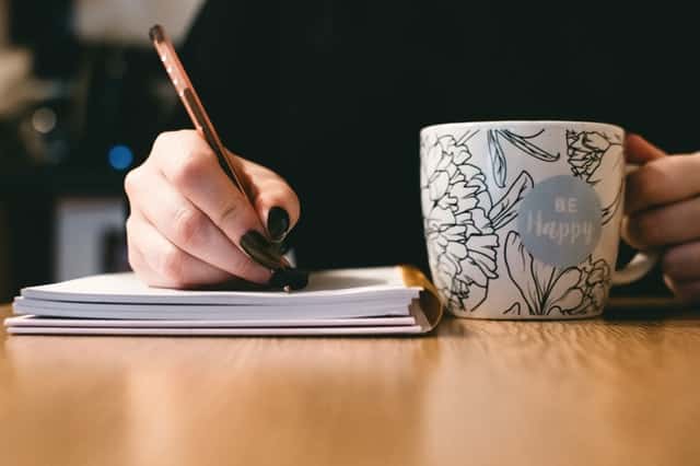 Mulher escrevendo em caderno com caneca ao lado