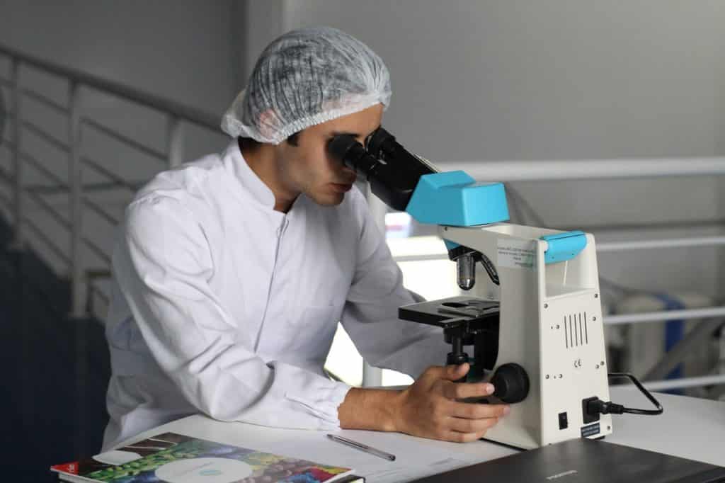 Profissional sentado com equipamentos de higiene olhando através de um microscópio.
