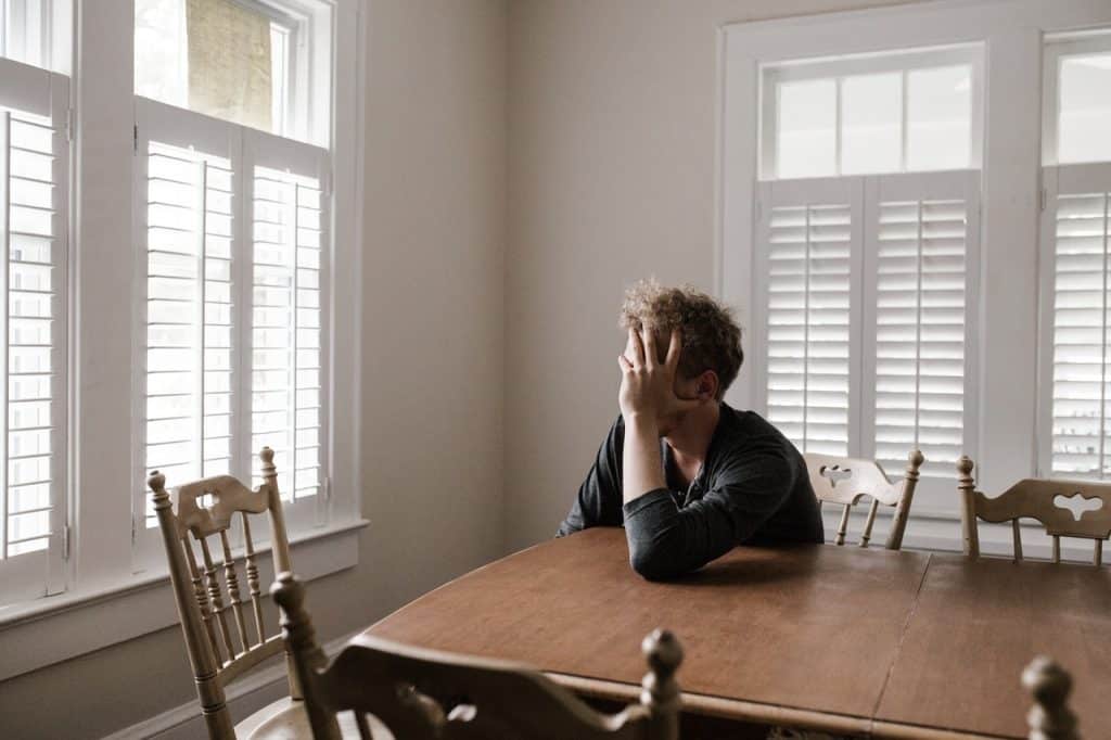 Homem sentado à mesa, em frente à janela, com as mãos segurando o rosto.