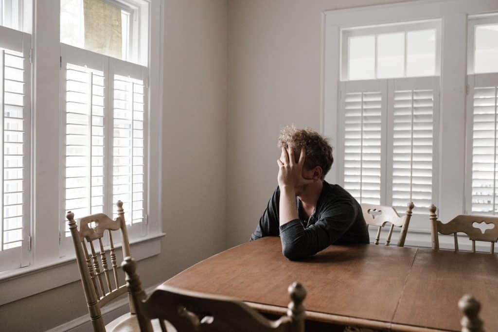 Foto de um homem sentado à mesa, sozinho, apoiando a cabeça em sua mão.