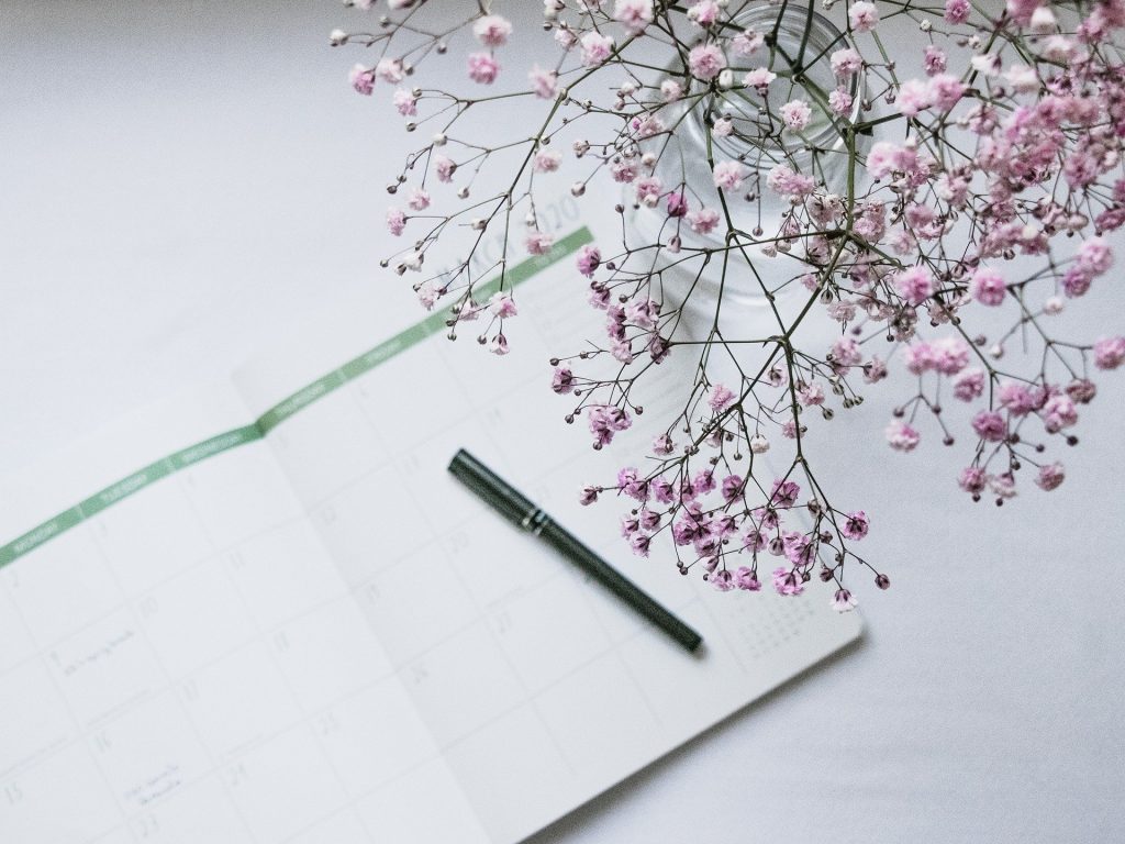 Imagem de uma agenda aberta e sobre ela uma caneta preta. Ao lado um vaso com lindas e pequenas flores rosas. 
