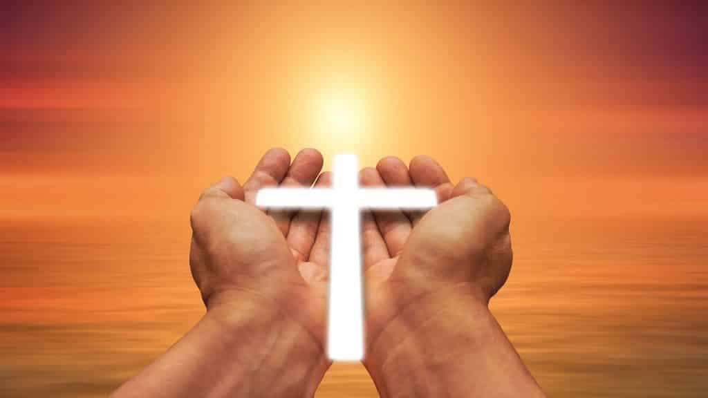 Imagem de duas mãos segurando uma pequena cruz iluminada. Ao fundo a imagem do sol bem alaranjado, representando a oração do perdão.