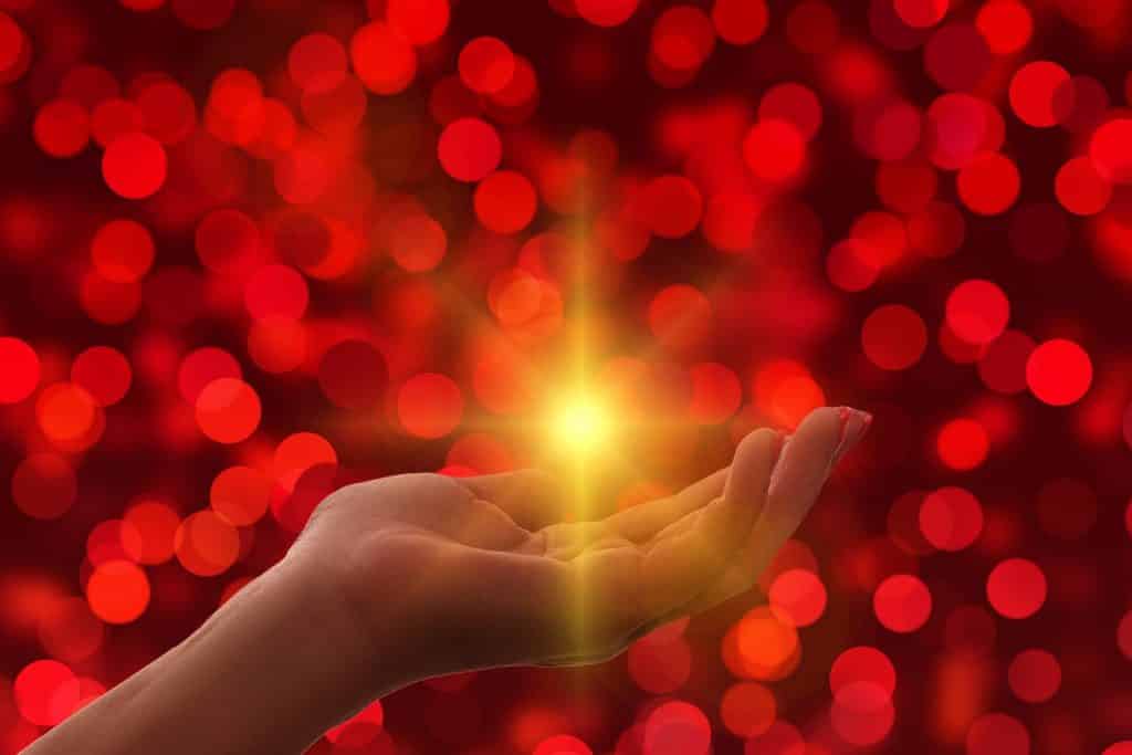 Imagem de uma mão feminina e ao centro uma luz amarela. A imagem tem um fundo iluminado com bolas vermelhas, representando a oração do perdão.