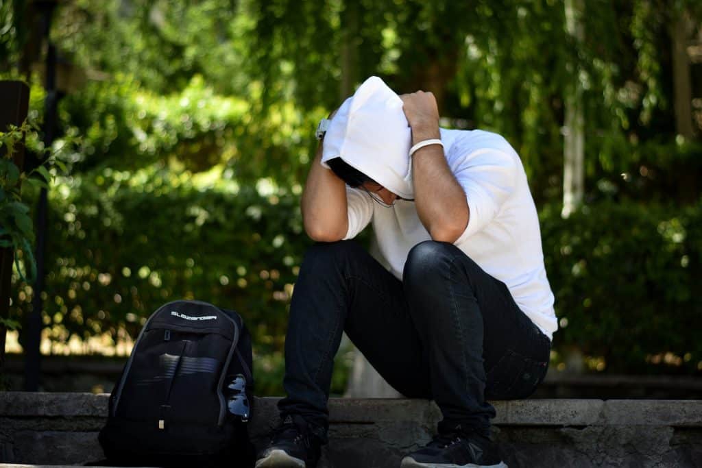 Imagem de um jovem sentado em um banco de praça. Ele está cabisbaixo e pensativo. Está com as mãos sobre a cabeça. Ao lado dele uma mochila preto. Parece que ele tem uma angústia no coração.
