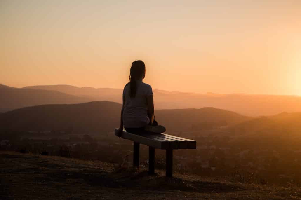 Mulher sentada  em um banco de madeira olhando para o pôr do sol