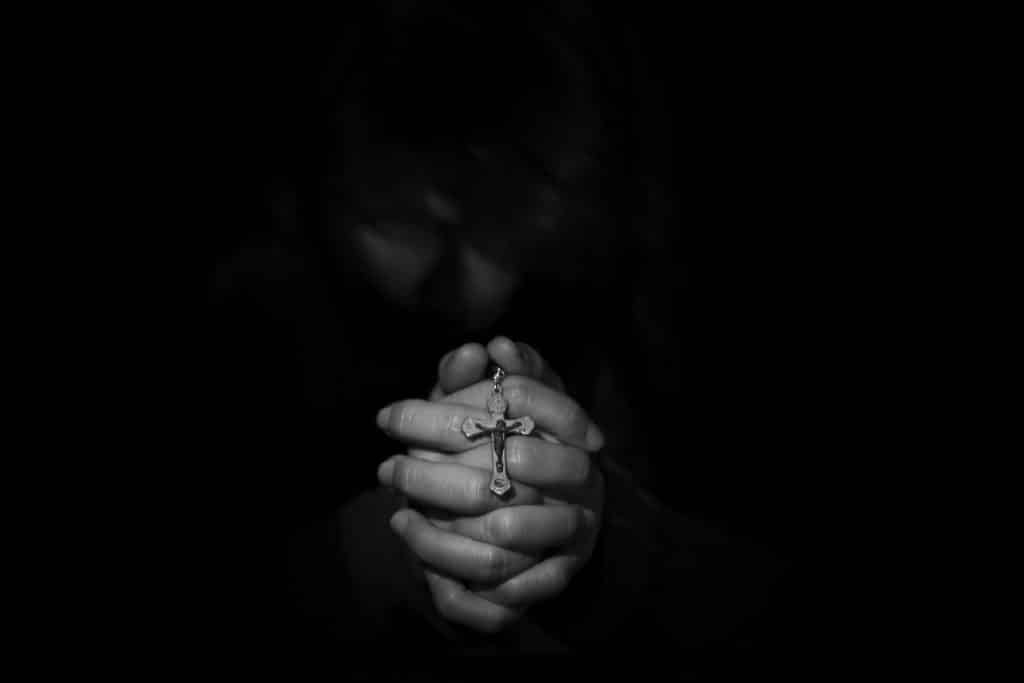 Imagem com fundo preto de uma mão feminina em posição de oração. Ela segura entre as mãos um crucifixo. Ela está fazendo a oração do perdão.