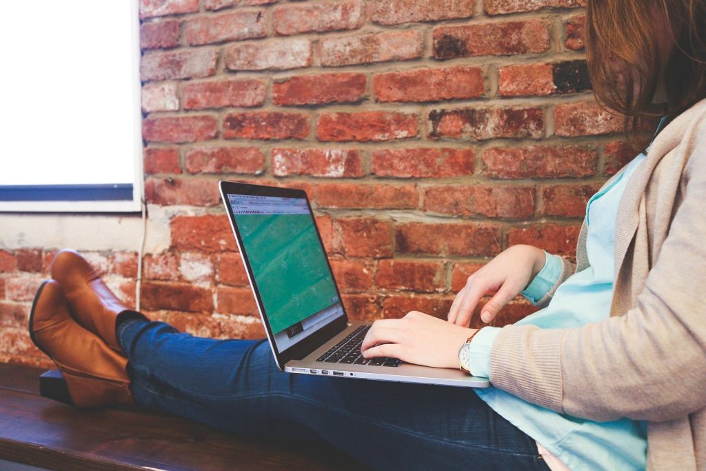 Imagem de uma mulher sentada sobre um banco de madeira e sobre o seu colo um notebook onde ela está fazendo planos para o futuro.
