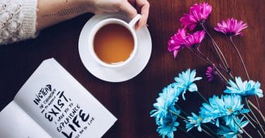 Mulher segurando xícara de chá na mesa ao lado de um livro e algumas flores